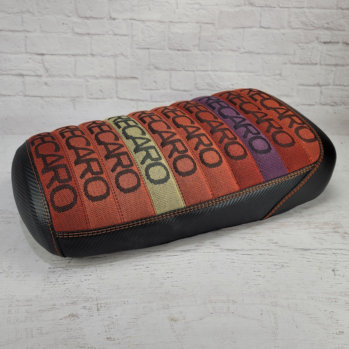 Front three-quarter view of the Recaro Sunrise Maddog seat cover showing repeating RECARO text in colorful sunrise stripes with orange stitching and carbon-fiber-look sides, handmade by Cheeky Seats