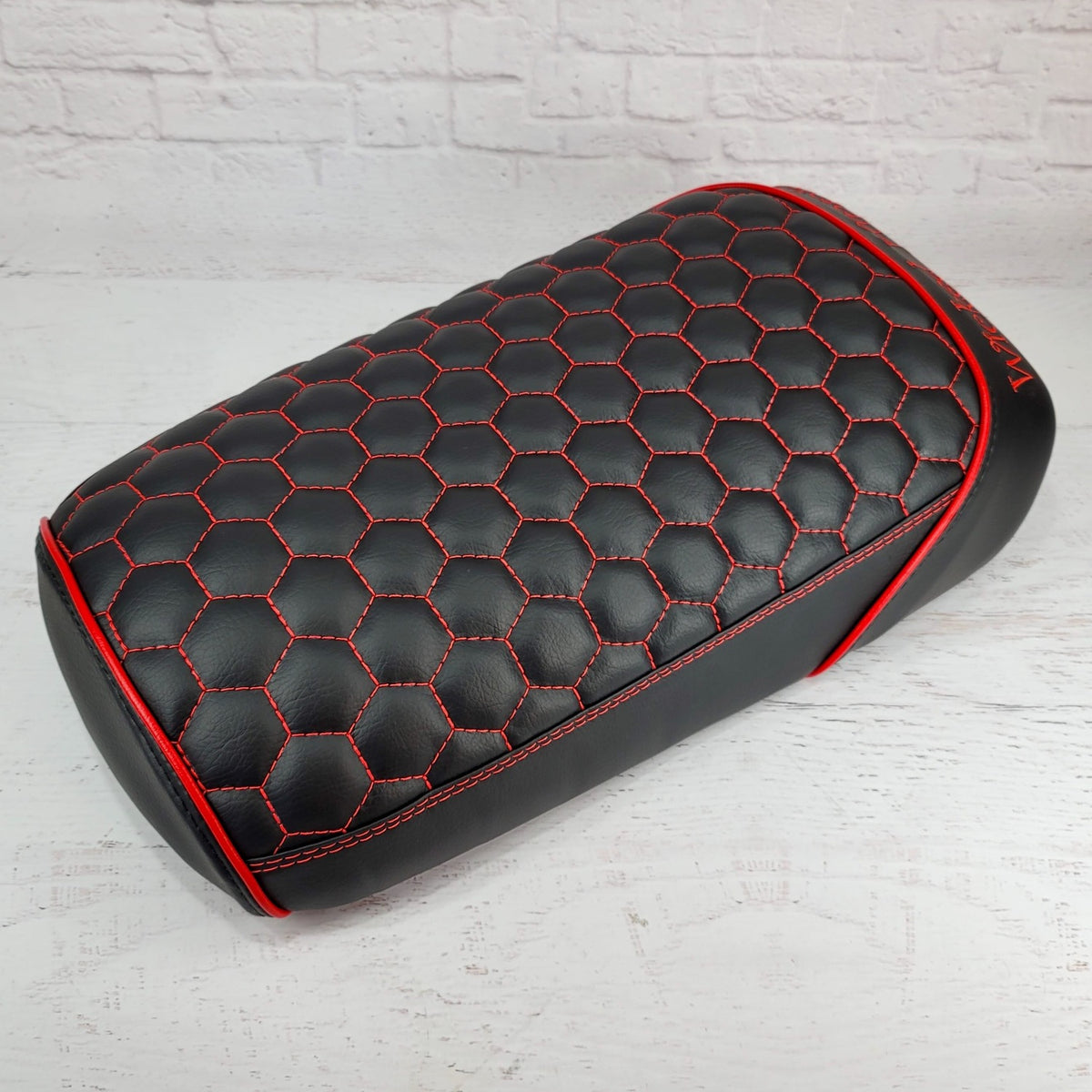 Black hexagon honeycomb-stitched Maddog seat cover with red stitching and red piping, three-quarter view against a white brick backdrop, handmade by Cheeky Seats