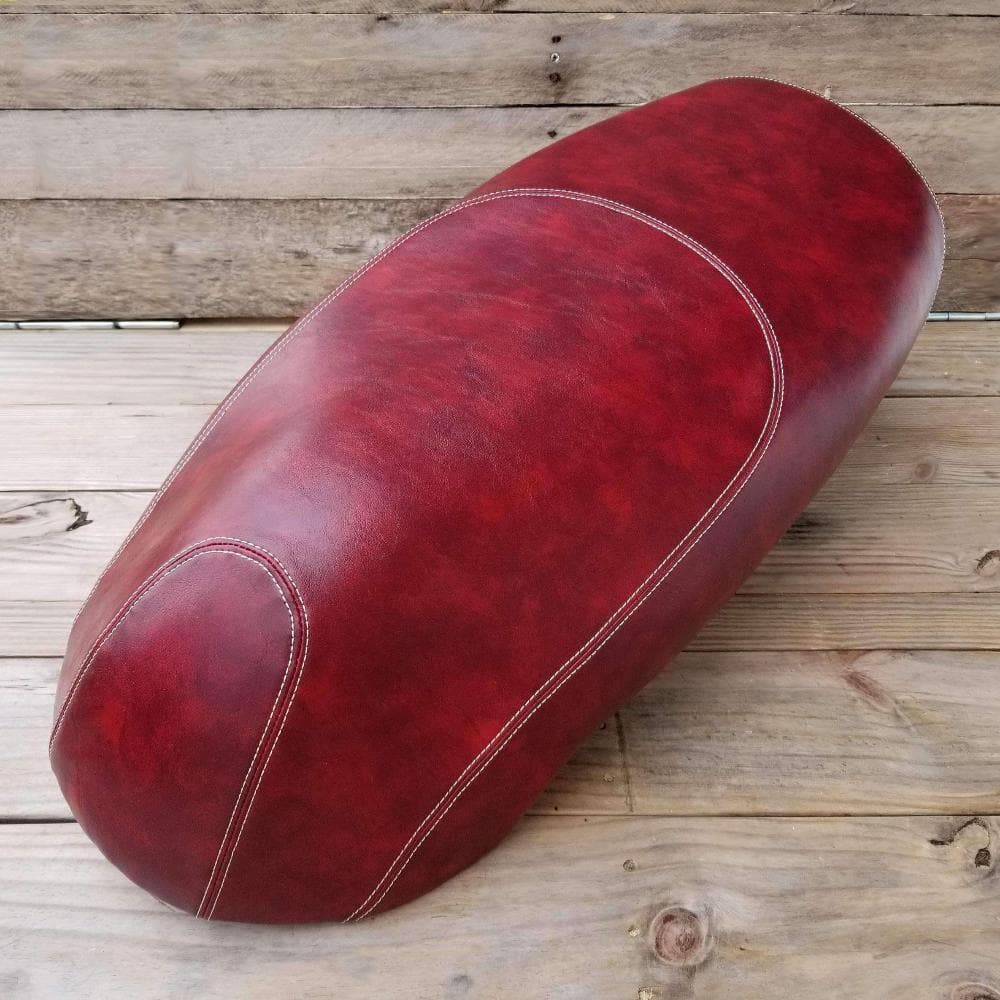 Rich mottled oxblood Kymco Like seat cover on weathered wood planks, overhead angle with cream contrast stitching along the center panel and side seams, handmade by Cheeky Seats